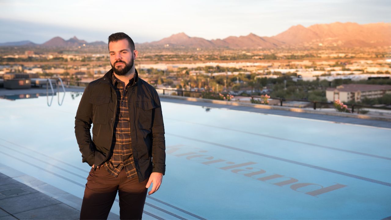 A man stands by an outdoor infinity pool with "Atierland" written in the water, overlooking a cityscape and mountains at sunset.